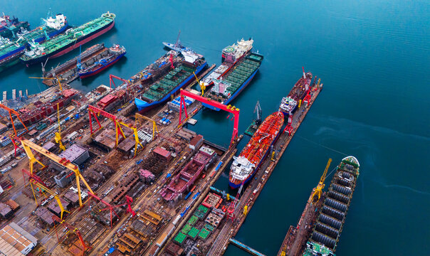 High angle aerial perspective of massive industrial shipbuilding complex showing heavy dry dock equipment red gantry cranes and large cargo vessels bordering calm ocean