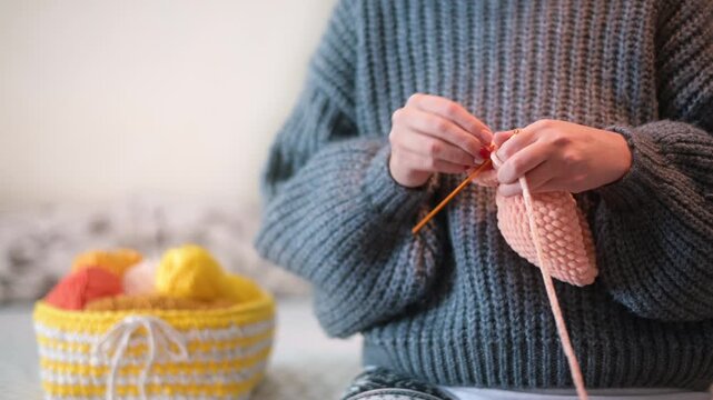 Close up of hands knitting pink yarn with basket of colorful yarn in background.
