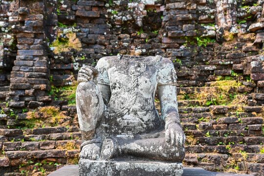 Ancient headless stone statue of a Hindu deity or guard in front of a red brick wall at My Son Sanctuary ruins in Vietnam