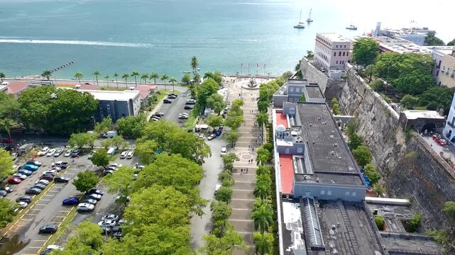 Drone footage of the Paseo la Princesa with a large fountain at the end, tourist walking and some sailboats at the bay of San Juan during the day.