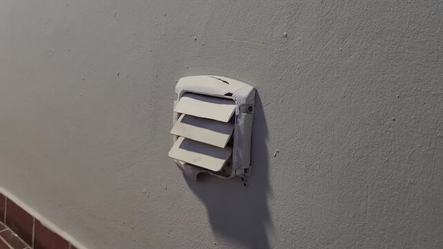 Detailed close-up of a cracked and deteriorating white wall ventilation cover attached to a textured exterior surface. Aging infrastructure and maintenance concept.