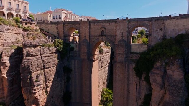 Drone footage rising and tilting down over Ronda's Puente Nuevo, framing the massive stone arches spanning the sheer gorge, with visitors lining the bridge railings above.