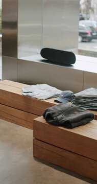 Folded casual garments on simple wood blocks. The shop interior is simple and modern with large windows. A skateboard leans on a ledge in the background. Cars pass on a city street outside.