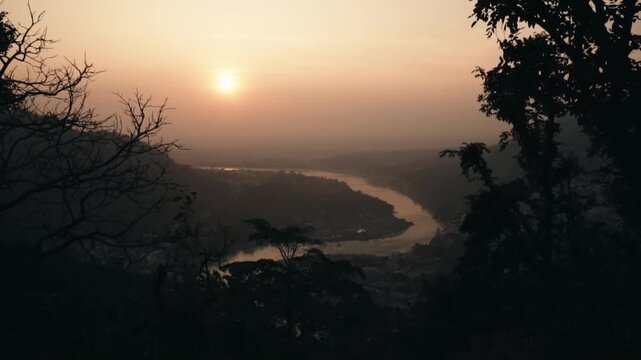 Breathtaking aerial view of a winding river flowing through a mountain valley with lush forest, illuminated by the warm golden light of the sun during a hazy and beautiful sunset
