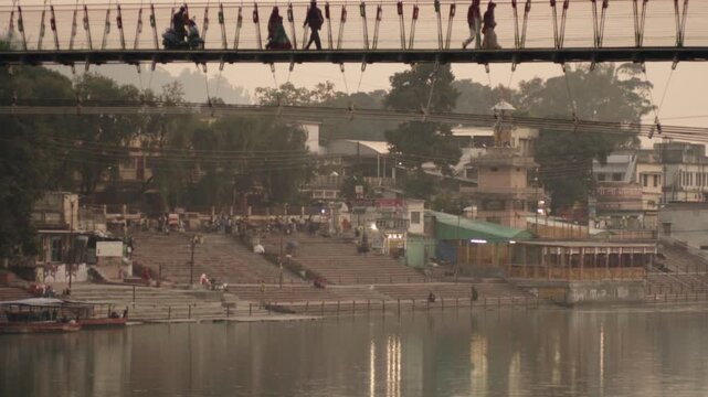 Local people and tourists crossing the famous Ram Jhula suspension bridge over the sacred Ganges river in Rishikesh, with the ghats visible on the far bank during a peaceful evening