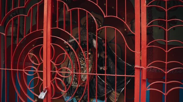 Ornate black statue of the Hindu goddess Kali adorned with garlands and jewels, enclosed behind a locked, decorative red metal gate at a vibrant traditional temple in India