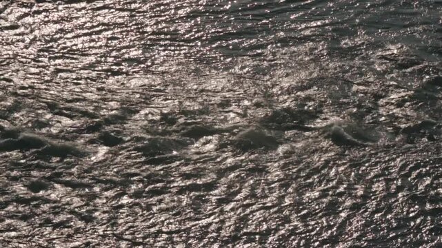Dark water surface moving restlessly with ripples and small waves, creating a mesmerizing shimmering light effect as it reflects the bright sunlight from a high angle view