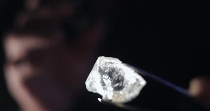 Slow motion examination of rough diamond held with tweezers in dark studio while jeweler inspects.