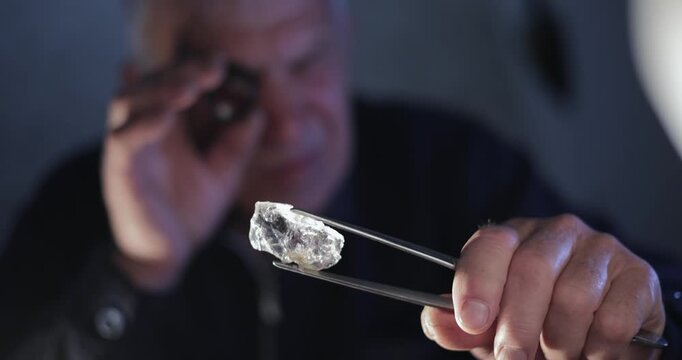 Slow motion man inspecting rough diamond with loupe and tweezers in gem lab.