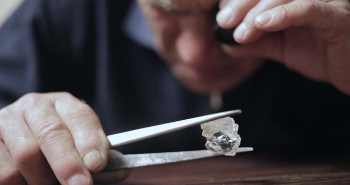 Closeup tracking shot of man inspecting rough diamond on wooden table in workshop using loupe and tweezers.