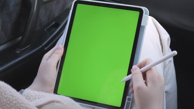 Person holds a tablet with green screen. They use stylus to draw or write. Focus remains on hands and device. Footage captures modern digital work in car. Ideal for apps, graphic design
