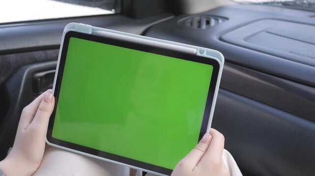 Hands hold tablet with green screen inside car. Person focuses on device, unaware of surroundings. Perfect for advertising, app demos, or digital content. Suitable for automotive, tech