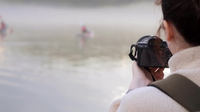 Photographer captures rowers on misty lake. Water reflects soft morning light. Person holds camera steady. Boats glide through calm surface. Mist rises gently above water