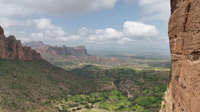 Breathtaking view from the Gheralta Mountains in Tigray, Ethiopia. Steep red sandstone cliffs overlook a vast, lush green valley with winding rivers and small farm settlements under a cloudy sky.