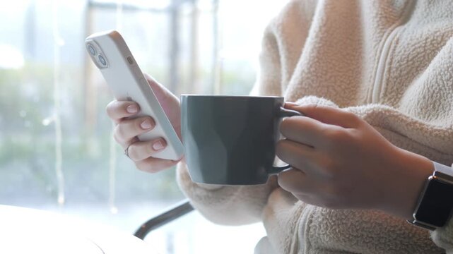 Person holds phone and coffee cup near window. Hands grip smartphone and dark mug. Person wears light-colored fleece jacket. Watch visible on left wrist. Background shows bright, blurred window view