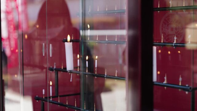 Person in dark jacket standing before rows of lit white votive candles inside a red lacquered shrine cabinet in Tokyo