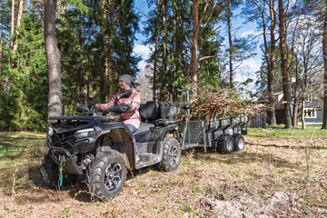 Fototapeta premium A woman rides an ATV quad with a trailer full of firewood along a green forest track in Estonia. She wears a jacket and hat while hauling branches and logs through trees.
