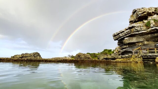 Scenic double rainbow in overcast sky, low view over seawater in rock pool