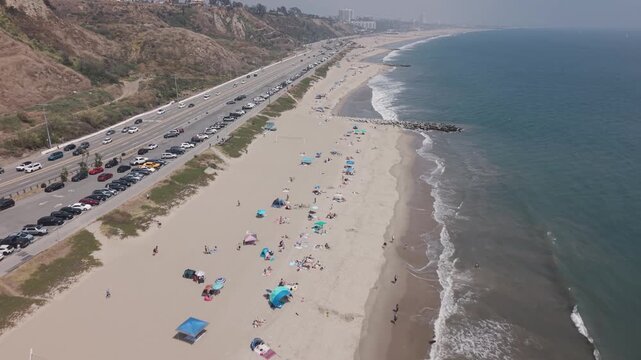 Will Rogers State Beach with beachgoers, colorful umbrellas, waves, busy Pacific Coast Highway traffic, rocky cliffs and distant coastline under hazy sky. California, USA. Aerial flyover shot.