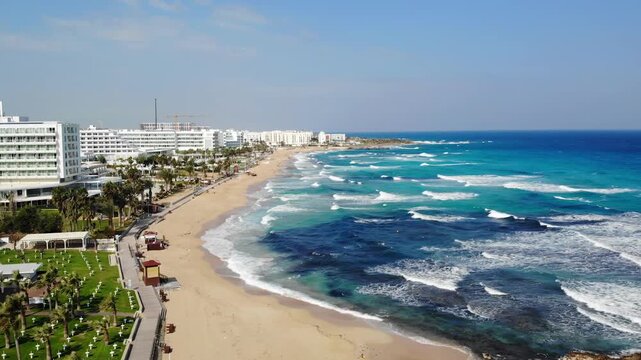 Aerial view of a beautiful sandy beach coastline on a sunny day, blue ocean waves washing ashore near hotels and palm trees.