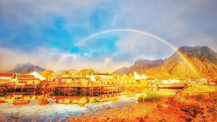 Svolvær waterfront cabins reflected on calm arctic water, with rainbow over the cloudy sky, Lofoten islands, Norway © Connie