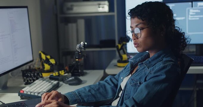 Young African American female programmer coding AI software on multiple screens in night office. Network security, cybersecurity, machine learning, working late, overtime, robotics automation concept.