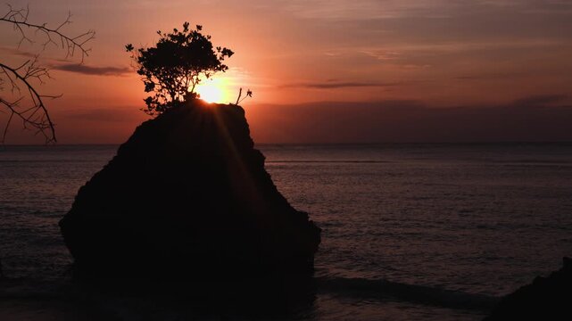 Padang Padang Beach (Pantai Labuan Sait) at sunset.  Island rock with tree in the foreground, with deep reds and purples of the setting sun in the background. The waves of the ocean can also be seen. 