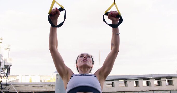 Bodyweight row. Urban fitness. Athlete female on rooftop performing physical exercise using suspension straps strengthening back biceps.