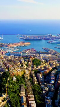 Vertical video. Genoa, Italy. Aerial panorama over Porto Antico, where dense terracotta rooftops meet the deep blue Ligurian Sea under bright summer light, with green hills rolling toward the horizon