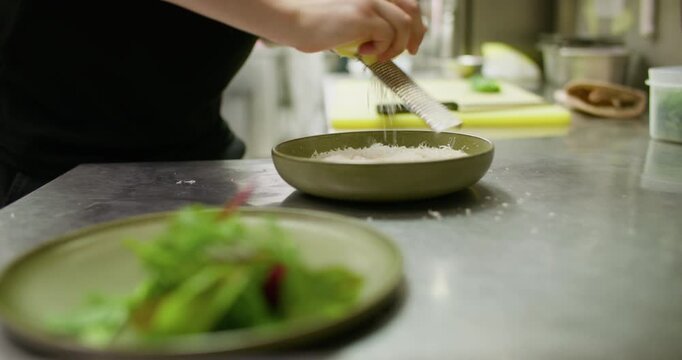 Shredding cheese. Food preparation. Unrecognizable professional chef man hands making presentation cooking parmesan salad dish on plate in restaurant.
