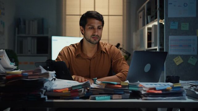 Overworked young Latin businessman working late on document in large stack of paperwork files at office night. Tax audit deadline, accounting stress, corporate burnout, urgent business concept.