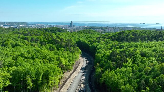 Cinematic aerial shot of a winding road through a lush green forest with the Gdynia city skyline and Baltic Sea in the background.