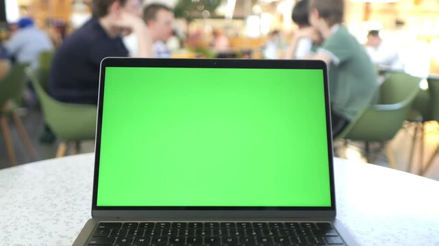 Laptop with green screen sits on table in cafe. Background shows blurred people seated at tables. Green screen ready for overlay use