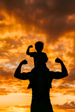 Silhouette of a strong father and son flexing muscles while the boy sits on his dad's shoulders against a dramatic sunset sky, superhero concept for Father's Day