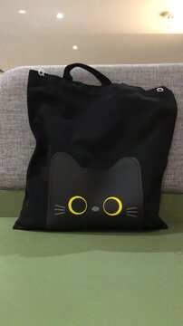A black tote bag with a cute black cat face design featuring large yellow eyes and whiskers, resting on a sofa.