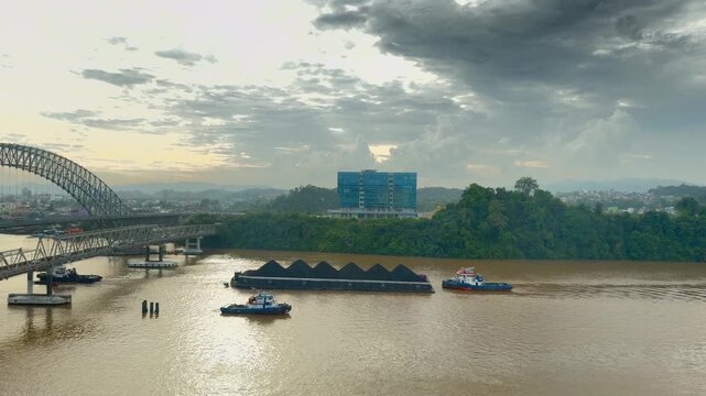 Large tugboats pull coal barges carrying fuel across the wide river near the massive steel arch Mahakam Bridge.