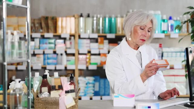 Mature female pharmacist standing behind the counter and working on computer, she checks the availability of medical medicines in a spreadsheet and prepares to collect an online order. High quality 4k
