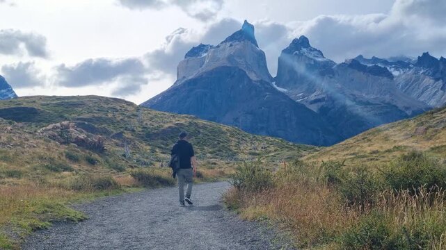 A lone traveler walks toward the iconic Cuernos del Paine peaks in Torres del Paine National Park, Chile, crossing vast Patagonian wilderness under dramatic cloudy skies