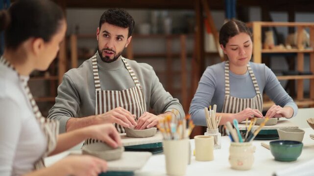 Two girl and guy craftspersons in striped aprons are busy molding clay products. They are sitting near work table, making plate out of raw clay. Working together in pottery workshop.