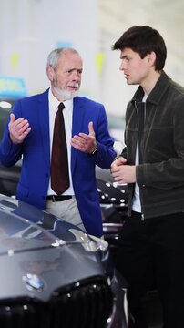 Mature male car seller in suit walking around car salon with guy who wants to buy a car and talking about specifications of cars. High quality 4k footage