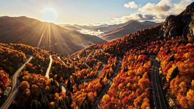 Aerial view of a winding serpentine mountain road through a vibrant autumn forest with red and orange foliage during a golden sunrise with bright sun rays and flare over the peaks.