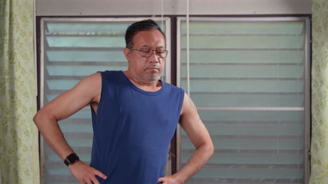 Serious mature Asian man wearing eyeglasses, navy sleeveless top, black smartwatch stands inside domestic room, keeping hands upon hips, looking sideways near glass louver windows, showing thoughtful.