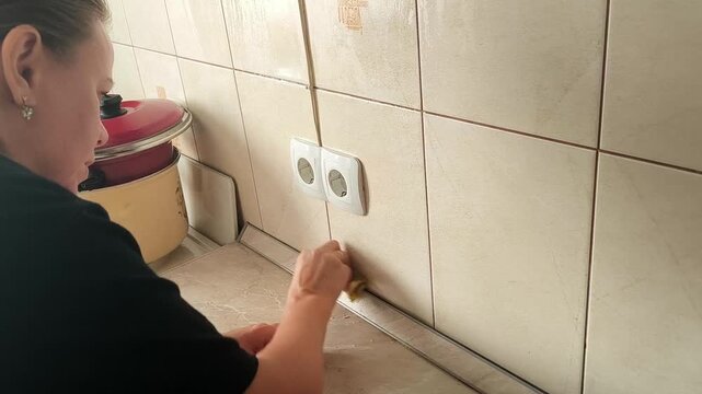 A woman wipes kitchen outlets and a tile apron next to a work area, a household cleaning and home care scene.
