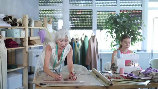 Woman creates wardrobe items, sews dresses and suits for clients, makes sweater out of knitwear. Mature female assistant drawing paper dress pattern by curves