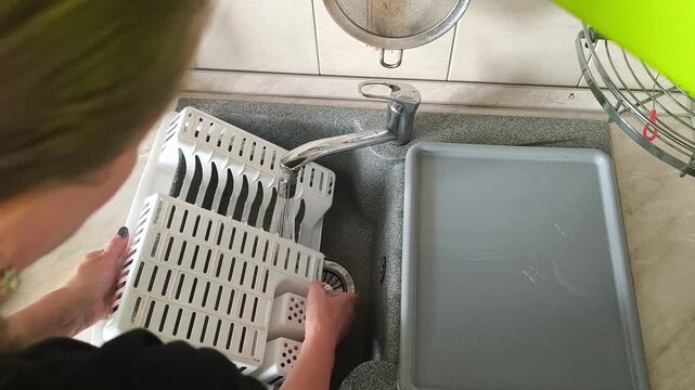 A woman rinses a plastic dish dryer under the faucet, a household scene of cleaning, kitchen maintenance and cleanliness.
