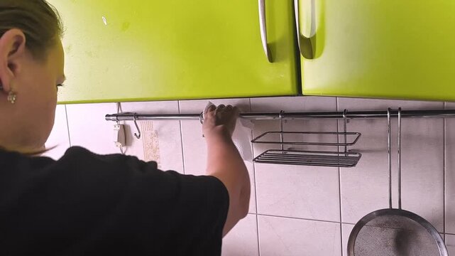 A woman cleans a metal kitchen railing with a napkin, a household scene of cleaning, kitchen maintenance and maintaining order.
