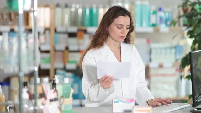 Processing of electronic document by computer. Woman employee in labcoat works in pharmacy using computer, remote document flow. Employee checks availability of drugs in pharmacy