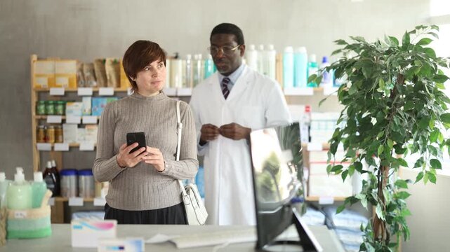 Woman in pharmacy with phone, looking for something in drug store. Client scrolls screen in search of electronic prescription. African American male pharmacist offer consultation in background