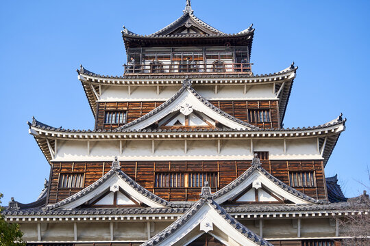 A close-up view of Hiroshima Castle's main keep (tenshu) against a clear blue sky, showcasing traditional Japanese feudal architecture. The multi-tiered structure features distinctive white plaster