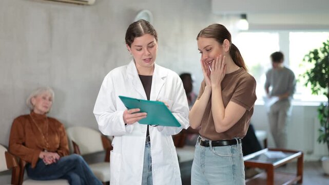 Serious nurse or doctor talking with young woman concerning documents while standing in reception area. High quality 4k footage